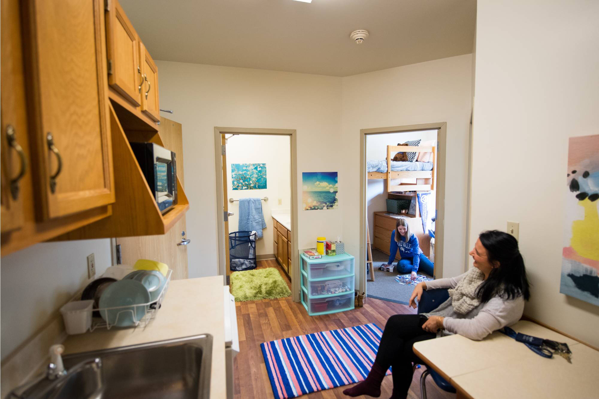 Cozy Johnson living unit with a kitchen, featuring wooden cabinets and a microwave. A woman sits at a table, smiling at a student in a doorway. It is a bright, cheerful vibe.